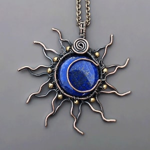 Może przedstawiać: Ręcznie robiony naszyjnik z wisiorkiem z motywem słońca i księżyca. Wisiorek ma ciemnoniebieski kamień lapis lazuli pośrodku, otoczony miedzianym drutem w kształcie słońca ze złotymi akcentami. Łańcuszek naszyjnika jest w kolorze brązu.