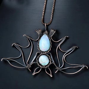 Pode incluir: Um colar com pingente de morcego feito à mão. O pingente apresenta um design de morcego feito de arame prateado, com duas pedras opalescentes em forma de pêra e redondas. A corrente do colar é de cor bronze escuro. O pingente é exibido em um fundo escuro.