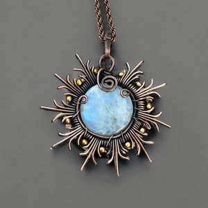 Op de afbeelding: Een handgemaakte koperen hangerketting met een ronde, lichtblauwe edelsteen in het midden. De edelsteen is omgeven door ingewikkeld koperdraadwerk in een zonnesterontwerp, geaccentueerd met kleine goudkleurige kralen. De ketting heeft een koperen ketting.
