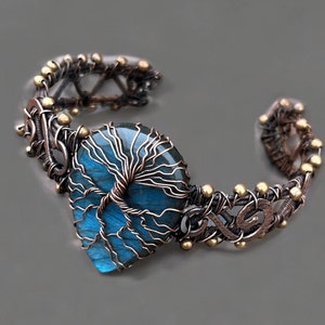 Puede incluir: Un brazalete artesanal de alambre de cobre con una piedra de labradorita azul en forma de corazón. La piedra está envuelta en alambre con un diseño de árbol de la vida. El brazalete tiene un estilo rústico y bohemio con detalles de latón.