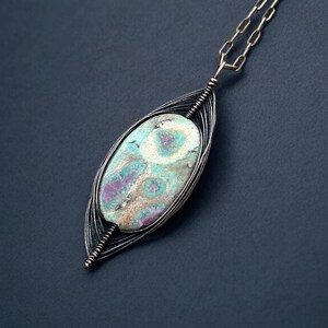 May include: A handcrafted pendant necklace featuring a large, oval-shaped stone with a unique pattern of teal, purple, and cream colors. The stone is encased in a silver-toned wire frame and suspended from a delicate chain.