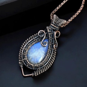 May include: A handcrafted pendant necklace featuring a teardrop-shaped, blue gemstone. The stone is encased in a woven, copper-colored wire frame, with decorative swirls and a matching chain. The background is dark.