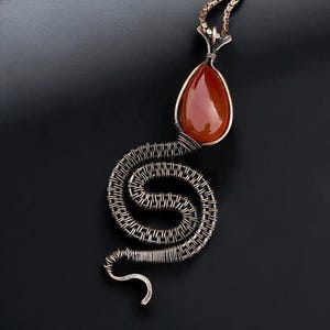 May include: A handcrafted pendant necklace featuring a teardrop-shaped, reddish-orange gemstone. The gemstone is set in a copper-colored metal frame and is connected to a coiled, wire-wrapped snake design. The necklace has a dark background.
