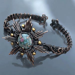 May include: A handcrafted copper bracelet with a central sunburst design. The focal point is a round, multi-colored stone, surrounded by radiating copper wires and small gold-colored accents. The bracelet has an open cuff design.