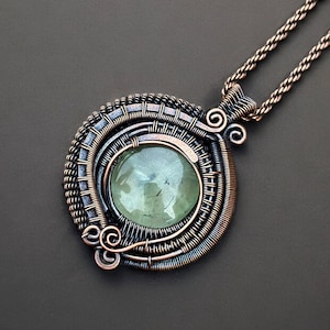 May include: A handcrafted pendant necklace featuring a large, round, pale green gemstone set within a detailed wire-wrapped frame. The frame is made of copper-colored and silver-toned wire, with decorative swirls and a matching chain.