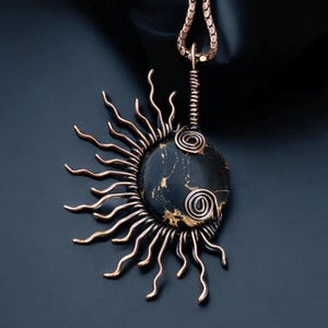 May include: A handcrafted pendant necklace featuring a black stone with gold veining, encircled by copper wire sun rays. The pendant hangs from a copper chain. The sunburst design is a unique piece of jewelry.