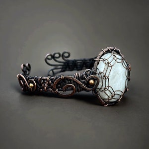 Puede incluir: Un brazalete artesanal de alambre de cobre con una piedra grande, de color claro y forma ovalada en el centro. El trabajo de alambre presenta patrones y espirales intrincados, con una pátina oscura en el metal. El brazalete está diseñado para usarse en la muñeca.