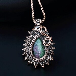 May include: A handcrafted copper pendant necklace featuring a teardrop-shaped gemstone with green and purple hues. The stone is encased in a woven wire setting with a sunburst design, and a decorative swirl. The necklace hangs from a delicate chain.