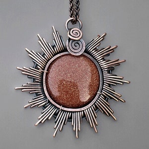 Puede incluir: Un collar con colgante de color cobre con un diseño de sol. El colgante tiene una piedra central, redonda, brillante y marrón. Los rayos del sol se extienden hacia afuera desde la piedra. Dos espirales adornan la parte superior del colgante.