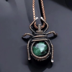 May include: A handcrafted pendant necklace featuring a wire-wrapped insect design. The pendant has a dark metal frame with copper wire detailing and a central green stone. The necklace chain is a copper color.
