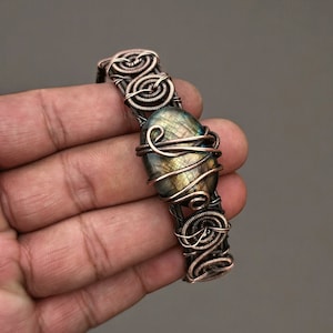 May include: A handcrafted copper wire bracelet with an oval gemstone at its centre, displaying iridescent blue and gold tones. The bracelet's design includes detailed wire wrapping and decorative spiral features, demonstrating a unique, artistic style.