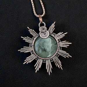 May include: A handcrafted copper pendant necklace featuring a sunburst design. The pendant has a central, round, green gemstone. The sunburst design has radiating lines. The necklace chain is a copper color.