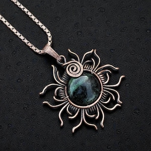 May include: A handcrafted pendant necklace featuring a dark green gemstone set within a copper-colored sunburst design. The pendant hangs from a silver chain, creating a striking contrast against the dark background. The necklace is a unique piece of jewelry.