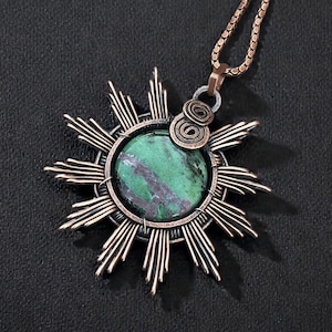 May include: A handcrafted pendant necklace featuring a green stone with dark purple veining, set within a copper-colored sunburst design. The pendant hangs from a matching chain, creating a unique and artistic accessory.