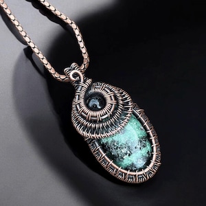 May include: A handcrafted pendant necklace featuring a green stone encased in a copper wire wrap. The pendant hangs from a matching copper chain. The stone has a speckled appearance, and the wirework is intricate.