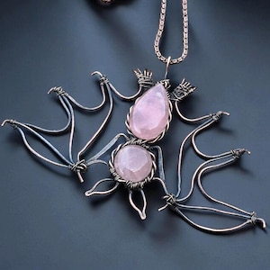 May include: A handcrafted bat-shaped pendant necklace. The pendant features a wire-wrapped design with two pink rose quartz stones. The wings are made of wire, and the necklace has a silver chain. The pendant is displayed on a dark background.