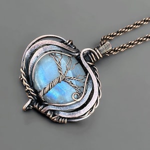 May include: A handcrafted pendant featuring a pear-shaped, iridescent moonstone. The stone is encased in a detailed, oxidized copper wire frame with a tree design. The pendant hangs from a matching copper chain, creating a unique, nature-inspired jewelry piece.
