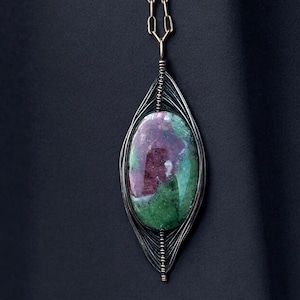 May include: A handcrafted pendant necklace featuring a large, oval-shaped gemstone with a mix of green and maroon hues. The stone is encased in a silver-toned wire frame, suspended from a delicate gold-colored chain. The design is elegant and artistic.