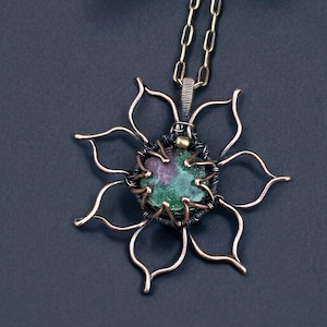 Pode incluir: Colar com pingente de cobre feito à mão com um design floral. O pingente apresenta uma pedra central com tons verdes e vermelhos, embutida em uma moldura de arame. As pétalas da flor são de cobre. A corrente do colar é dourada.