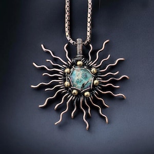May include: A handcrafted pendant necklace featuring a sunburst design. The pendant has a central, multi-faceted, teal-colored stone, surrounded by copper-colored rays and small gold-colored beads. The necklace chain is a silver-colored, textured link.