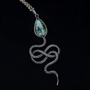 May include: A pendant necklace featuring a teardrop-shaped green and magenta stone set in a wire-wrapped metal frame shaped like a coiled snake. The metal wire has a dark, oxidized finish and connects to a matching silver-toned chain link necklace.