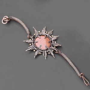 May include: A handcrafted bracelet featuring a sunburst design. The central element is a round, pinkish-orange stone, surrounded by silver-toned wire rays and small gold-colored beads. The bracelet has a textured, copper-colored chain.