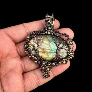 May include: Oval pendant featuring a labradorite stone with a tree design, encased in copper wire with brass accents. The pendant has a detailed, handcrafted appearance, showcasing a natural landscape within the stone.