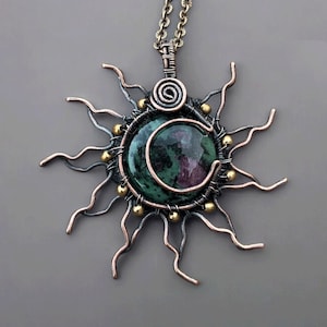 May include: A handcrafted pendant necklace featuring a sunburst design. The pendant has a central, round, green and red stone, encircled by wire and accented with gold-colored beads. The sun's rays are made of wavy copper-colored wire.