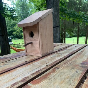 May include: A wooden birdhouse with a small entrance hole and a perch. The birdhouse is sitting on a weathered wooden table.