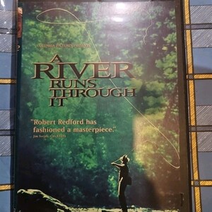 May include: DVD case for "A River Runs Through It" with a green and brown forest background. The title is in large gold letters. A silhouette of a person fly fishing is in the lower right corner. The case also includes a quote from Jim Svejda, CBS Radio.