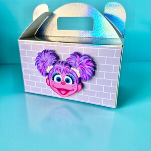 May include: A small, light purple gift box with a brick pattern and a holographic handle. The box features a colorful cartoon character face with purple hair and a pink mouth. The box is on a light blue surface.