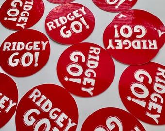 Puede incluir: Un conjunto de pegatinas rojas y circulares con el texto blanco "RIDGEY GO!". Las pegatinas están dispuestas sobre una superficie blanca. El diseño presenta una fuente en negrita y un signo de exclamación.