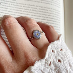 Op de afbeelding: Een zilveren ring met een delicaat kettingontwerp heeft een blauwe vergeet-mij-nietje bloem geperst in een heldere ovale harssetting.