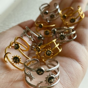 Puede incluir: Una colección de anillos con temática celestial en tonos dorados y plateados. Los anillos presentan diseños de sol, luna y estrellas con centros oscuros. Algunos anillos tienen bandas abiertas, mientras que otros son sólidos. Los anillos se muestran en una mano.