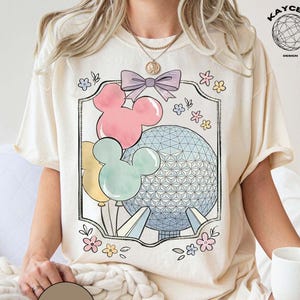 May include: Cream-colored t-shirt featuring a framed graphic with Mickey Mouse balloons in pink, yellow, and mint green, and a geometric sphere. The design includes a purple bow and floral accents. The word "NATURAL" is printed on a brown circle.