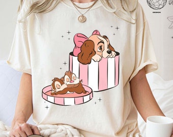 Puede incluir: Una camiseta blanca con una caja de rayas rosas y blancas con una imagen de dibujos animados de La Dama y el Vagabundo. Lady está en la caja y Tramp está fuera de la caja. El texto "NATURAL" está en la parte inferior de la imagen.