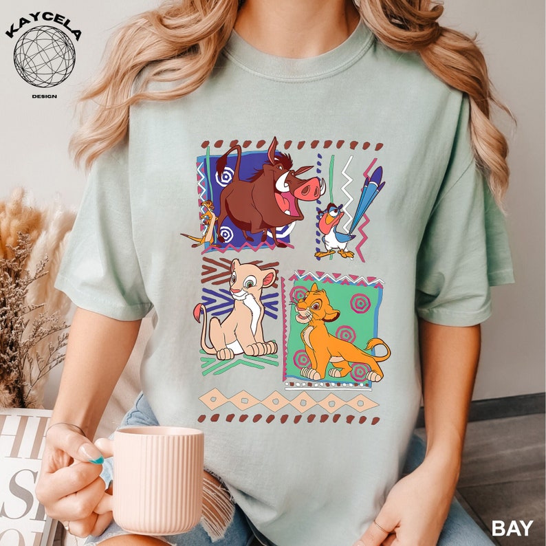 May include: A light blue t-shirt with a graphic print of characters from the movie The Lion King. The characters are Simba, Nala, Pumbaa, and Zazu. The graphic is surrounded by a colorful geometric pattern.