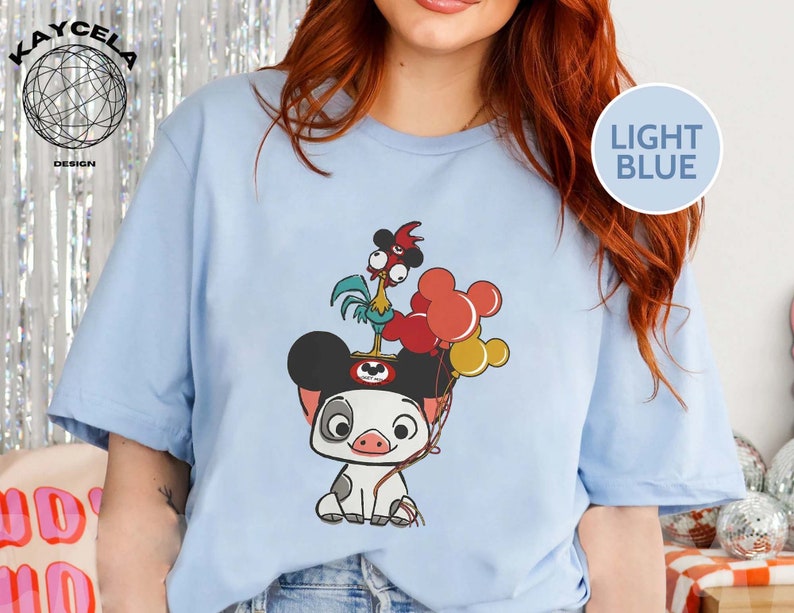 May include: Light blue t-shirt featuring a cartoon pig wearing Mickey Mouse ears and holding balloons. A rooster with a red comb is perched on top of the pig's head.