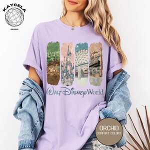 Op de afbeelding: Een lichtpaars t-shirt met een afbeelding van Walt Disney World-bezienswaardigheden. Het ontwerp bevat afbeeldingen van een kasteel, een boom en andere parkkenmerken. De tekst "Walt Disney World" staat onder de afbeeldingen. Het shirt heeft ook de tekst "Orchid Comfort Colors".