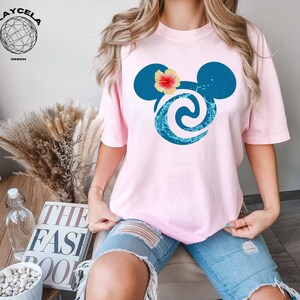 May include: Pink t-shirt featuring a blue and turquoise Mickey Mouse head design. The Mickey Mouse head has a pink flower on top.