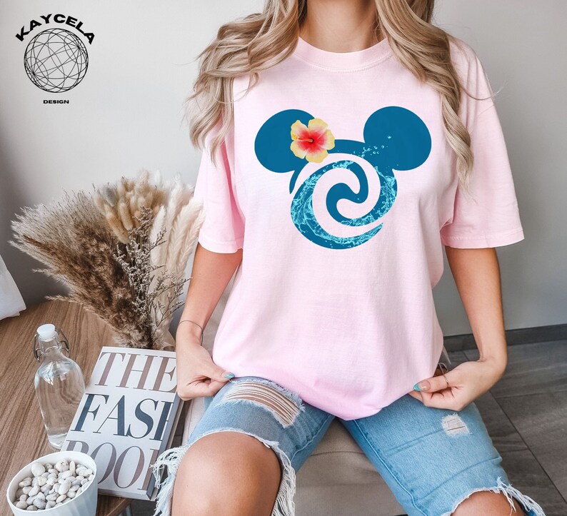 May include: Pink t-shirt featuring a blue and turquoise Mickey Mouse head design. The Mickey Mouse head has a pink flower on top.