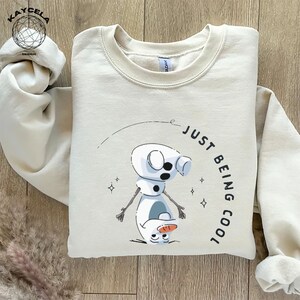 May include: A beige sweatshirt with a cartoon image of Olaf from Frozen. The text "Just Being Cool" is written in a circle around Olaf.