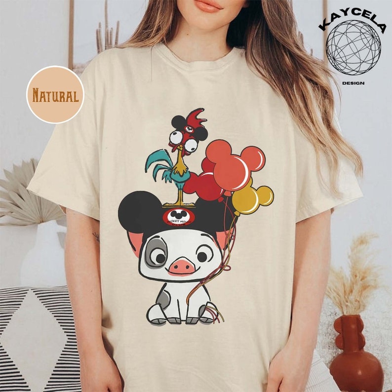 May include: A beige t-shirt featuring a cartoon pig wearing Mickey Mouse ears and holding balloons. The pig is wearing a red and white shirt with the words "Mickey Mouse" on it. A rooster with a red comb and blue and green feathers is standing on top of the Mickey Mouse ears.