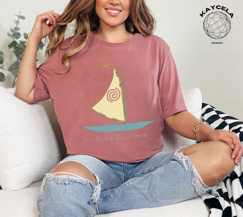 May include: A woman wearing a maroon t-shirt with a cartoon image of a sailboat and the text "it calls me". She is sitting on a white couch with a black and white checkered throw blanket.