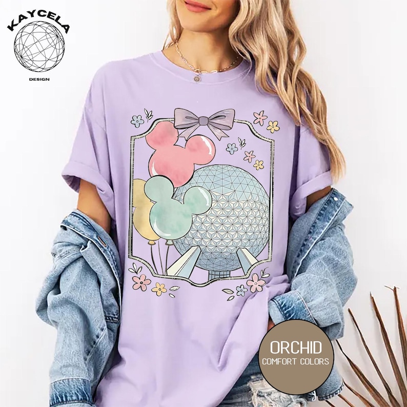May include: A light purple t-shirt featuring a graphic of Mickey Mouse balloons and a geometric sphere, with a bow and floral accents. The shirt has the text "ORCHID COMFORT COLORS" in a brown circle.
