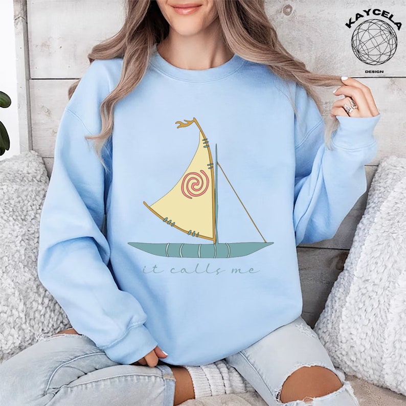 May include: A light blue sweatshirt with a cartoon image of a sailboat with a spiral design on the sail. The text "it calls me" is printed below the sailboat.