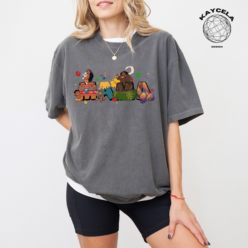 K&ouml;nnte beinhalten: Ein graues T-Shirt mit einem Aufdruck der Figuren aus dem Film Vaiana, die "MAUI" in bunten Buchstaben buchstabieren. Die Figuren tragen traditionelle hawaiianische Kleidung und sind von tropischen Pflanzen und Blumen umgeben.