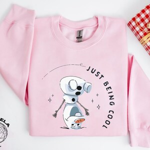 May include: Pink sweatshirt with a cartoon snowman graphic and the text "Just Being Cool".