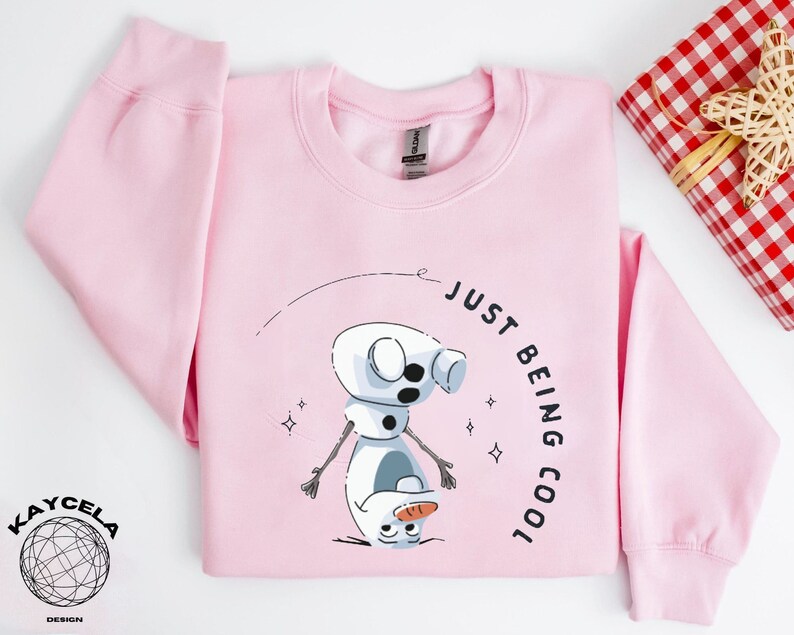 May include: Pink sweatshirt with a cartoon snowman graphic and the text "Just Being Cool".