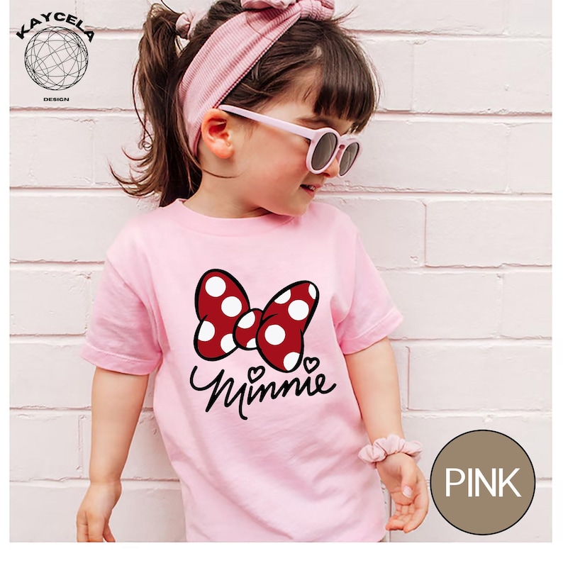 May include: A pink t-shirt with a red and white polka dot bow and the word "Minnie" written in black script. The shirt is being worn by a young girl with brown hair and pink sunglasses.